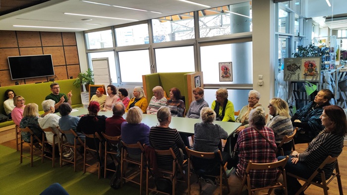 Grupa ok. 20 seniorek siedzi wokół stołu konferencyjnego i słucha z uwagą prowadzącego spotkanie.